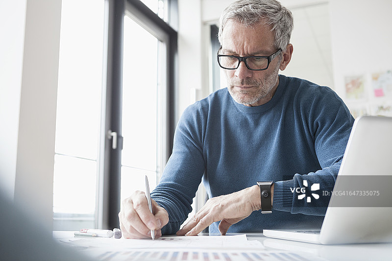 一个成熟的男人坐在办公室里用笔记本电脑和智能手表工作图片素材
