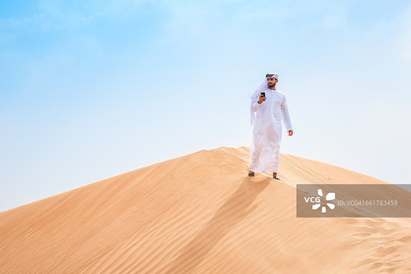 阿拉伯联合酋长国迪拜，一名身穿传统服装的中东男子在沙漠沙丘上使用智能手机图片素材
