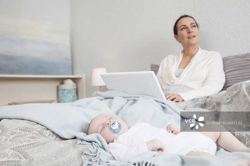 母亲坐在床上使用笔记本电脑，而男婴睡在她旁边图片素材