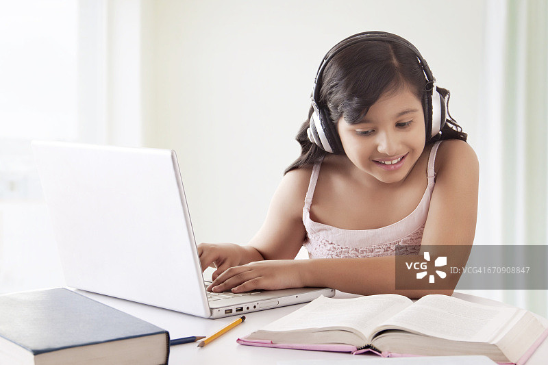 微笑的可爱女孩戴着耳机使用笔记本电脑和看教科书的肖像图片素材