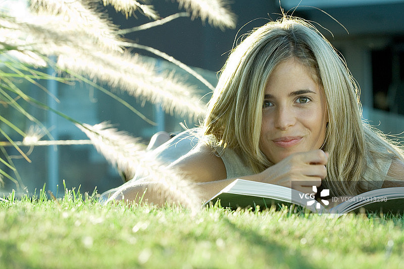 女人躺在草地上，看书，对着镜头微笑图片素材