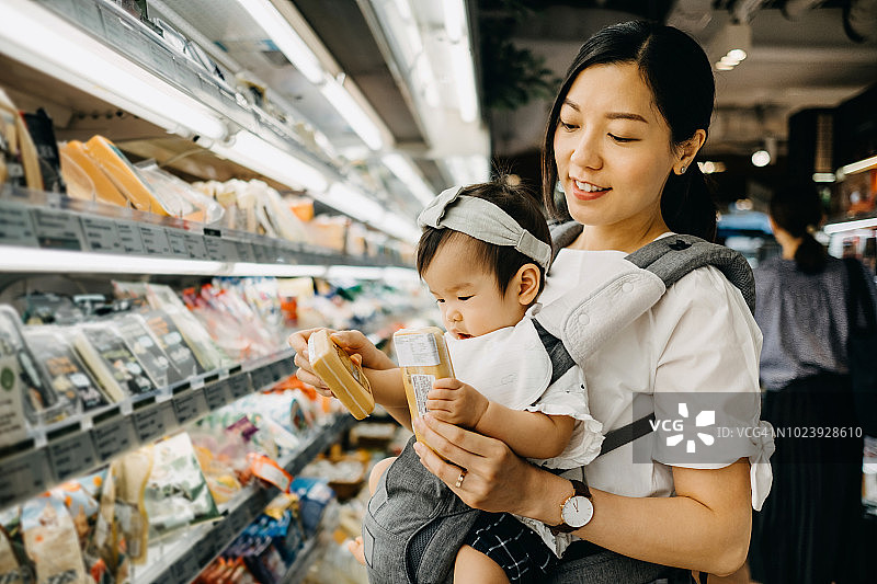 美丽的母亲抱着可爱的女婴在超市购物，阅读新鲜奶酪包装袋上的营养标签图片素材