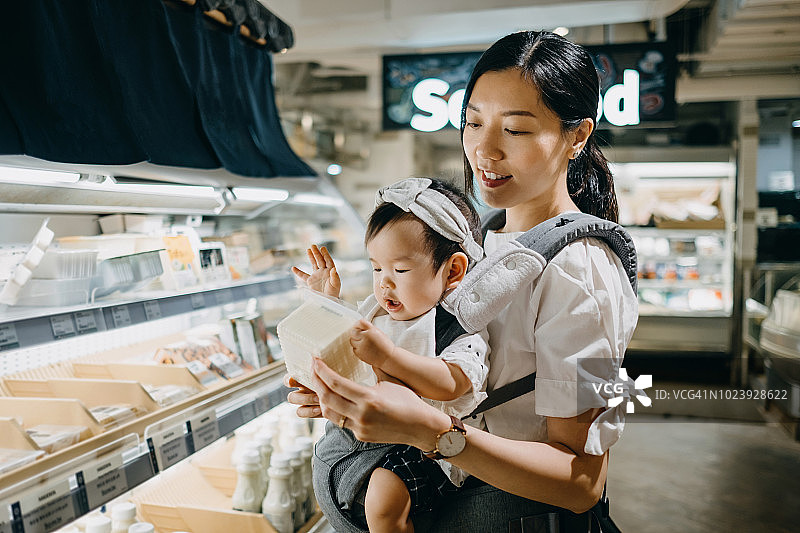 美丽的母亲抱着可爱的女婴在超市购物，阅读一包新鲜有机豆腐上的营养标签图片素材
