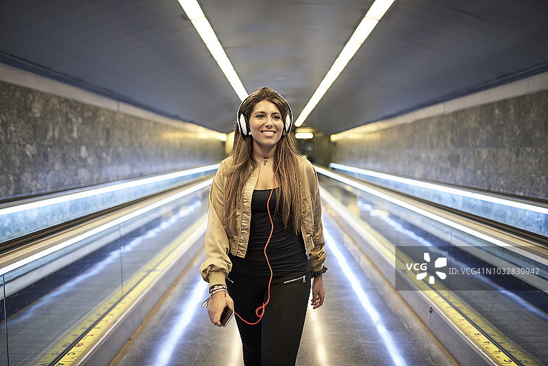 微笑的女人戴着耳机和智能手机在地铁站的肖像图片素材