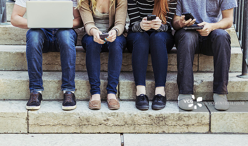 四名学生在大学校园的一个台阶上坐成一排，使用他们的技术，只能看到他们的脚和腿图片素材