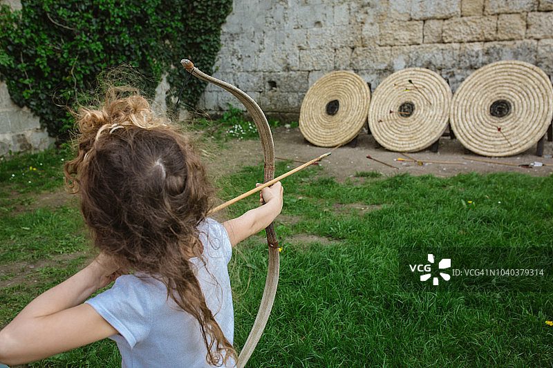 女孩手持中世纪木制弓箭，瞄准石头城堡前的三个干草靶子图片素材