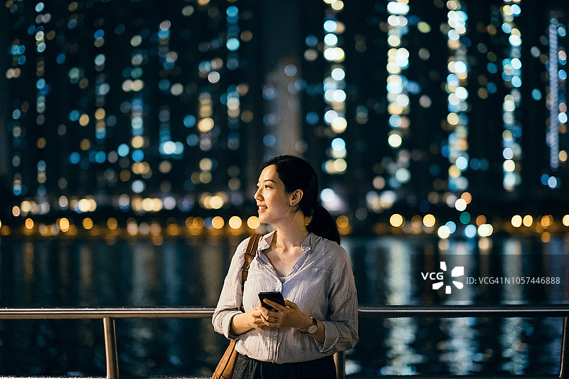 一名年轻女子晚上在港口边使用智能手机，与灯光照亮的城市景观形成对比图片素材