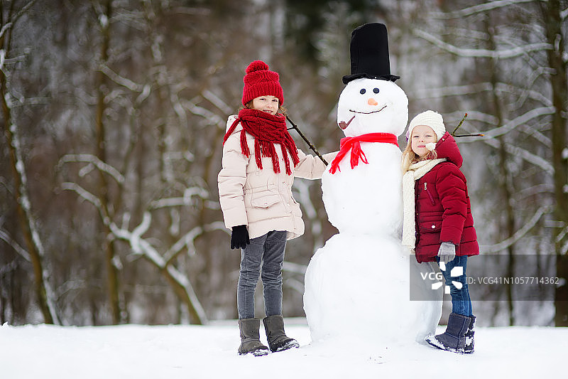 两个可爱的小女孩在美丽的冬季公园里一起堆雪人。可爱的姐妹们在雪地里玩耍。图片素材