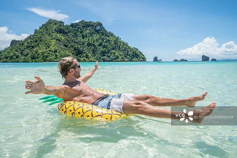 快乐的年轻人与充气菠萝漂浮在美丽的热带海滩享受海滩假期天堂。男性为了幸福和活力而伸出双臂图片素材