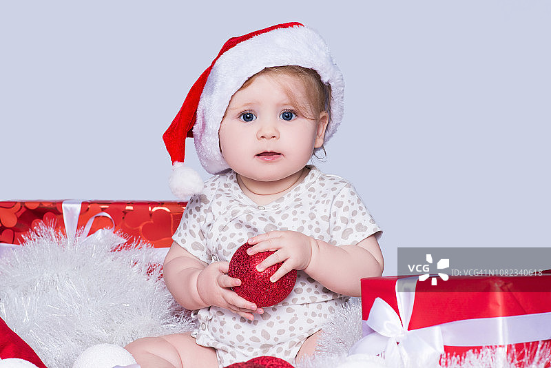 小女孩，戴着圣诞老人的红帽子，周围是圣诞球、礼盒和花环，在灰色的天空中孤立无存图片素材
