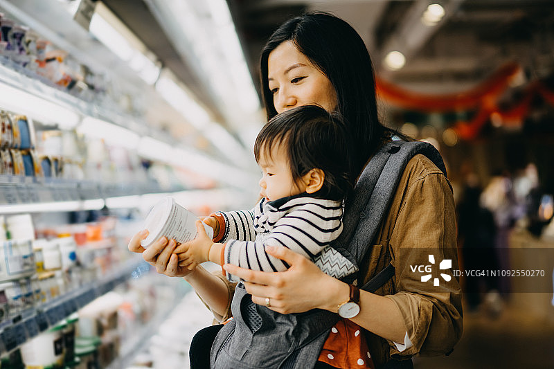 母亲抱着可爱的女婴在超市购物，阅读新鲜酸奶包装上的营养标签图片素材