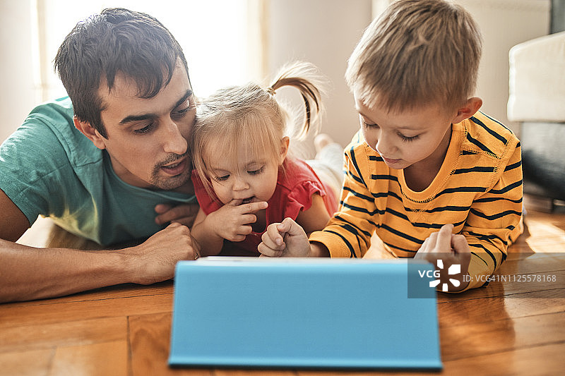 父亲和他的孩子们躺在家里的地板上使用数码平板电脑图片素材
