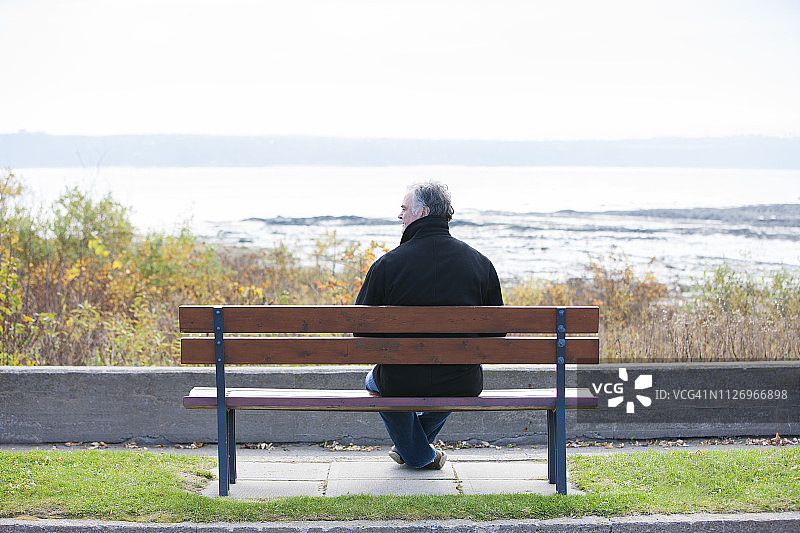 一个成熟的男人坐在公园长椅上俯瞰着河流。图片素材