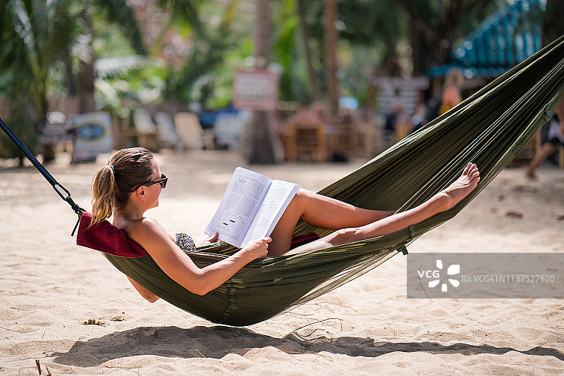 放松的女人在沙滩上的吊床上看书。图片素材