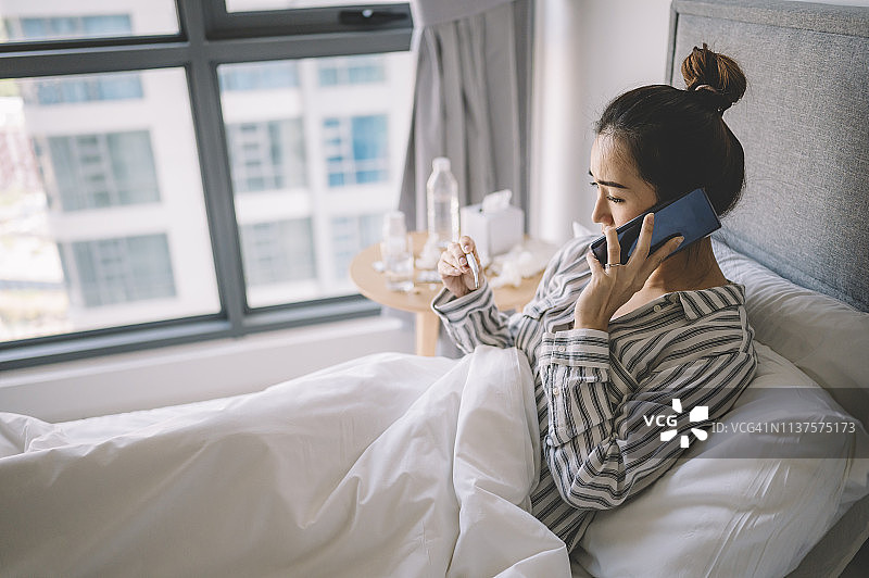 一位亚洲华人女性穿着睡衣在卧室边看床上的温度计边讲电话图片素材
