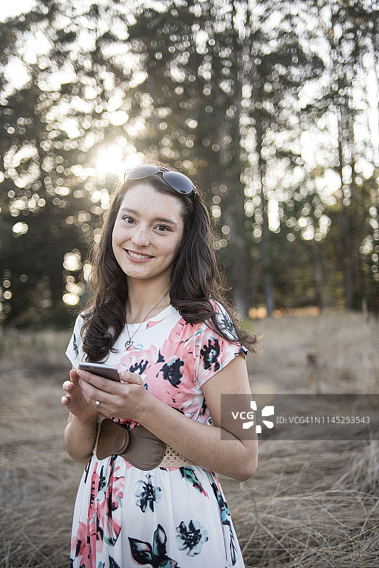 微笑的年轻女子的肖像使用手机，而站在森林的树木图片素材