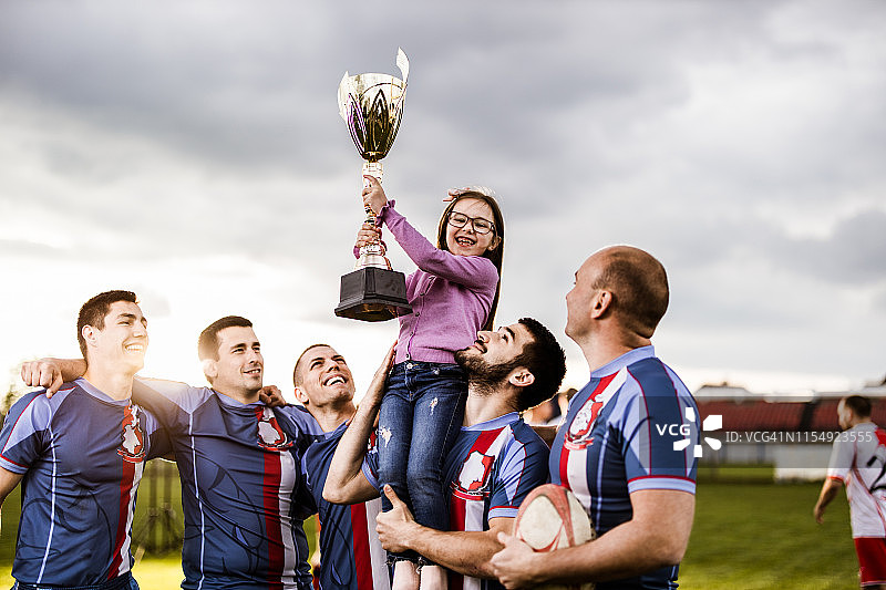 快乐的女孩和橄榄球队在操场上庆祝奖杯。图片素材