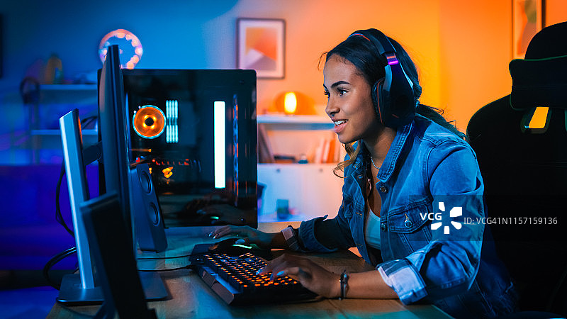 戴着耳机的漂亮和兴奋的黑人玩家女孩正在她的电脑上玩第一人称射击在线视频游戏。房间和电脑都有彩色霓虹灯。温馨的家庭之夜。图片素材