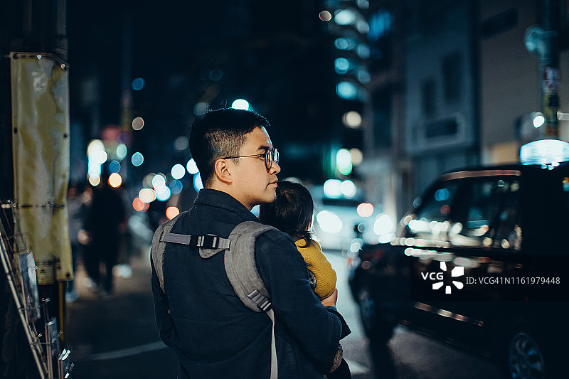 年轻的亚洲父亲抱着可爱的小女儿在夜晚穿过城市的街道，与繁忙的交通灯光景观图片素材
