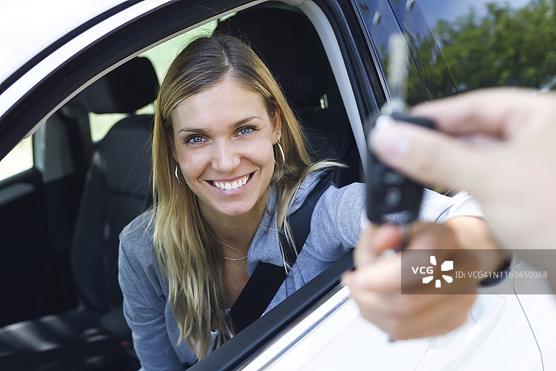 一幅微笑的年轻女子拿着车钥匙看着相机的照片，然后把钥匙交给窗外的人图片素材