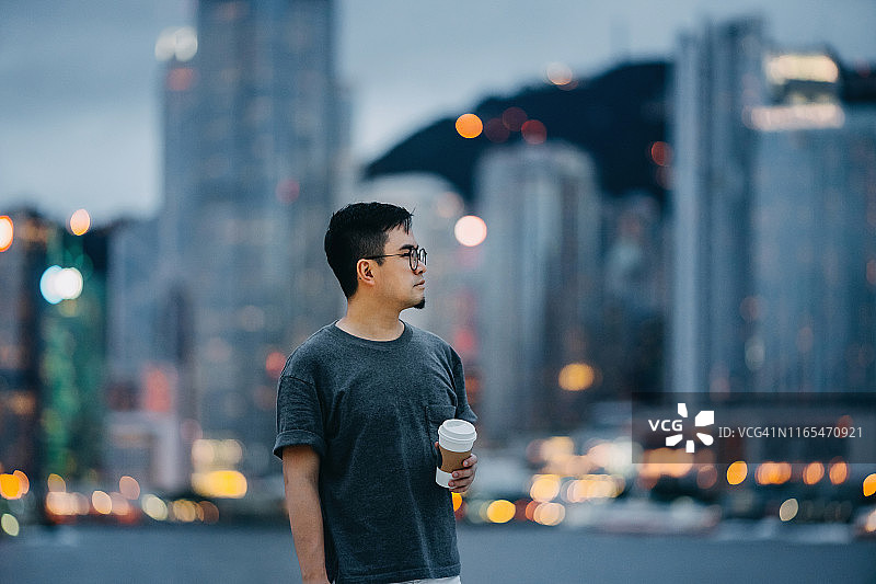 黄昏时分，在维多利亚港的海滨长廊上，聪明的年轻亚洲男子一边喝着咖啡，一边站在壮观的城市天际线前图片素材