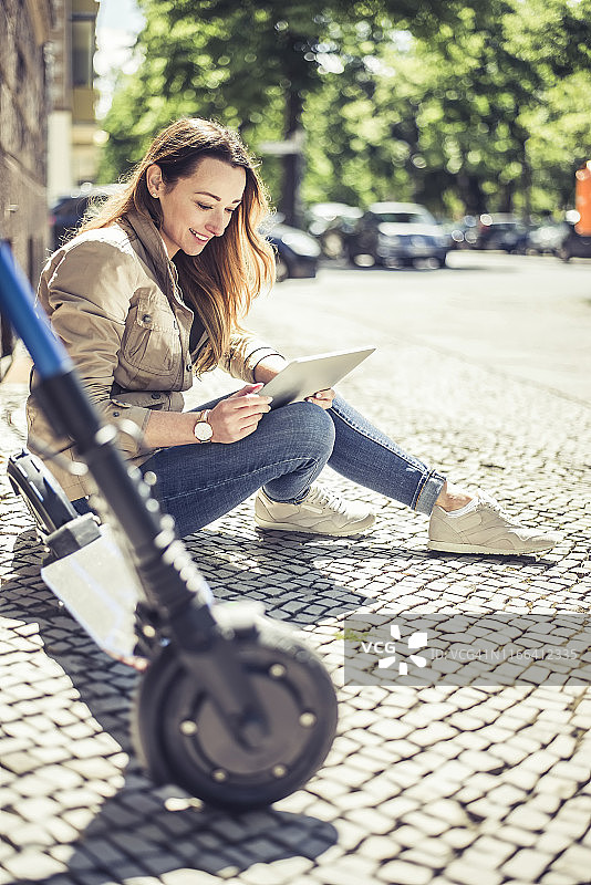 面带微笑的女人坐在人行道上，旁边是她的电动滑板车，她在用平板电脑图片素材