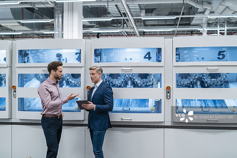 在一个现代化的工厂里，两个商人用平板电脑对着机器说话图片素材
