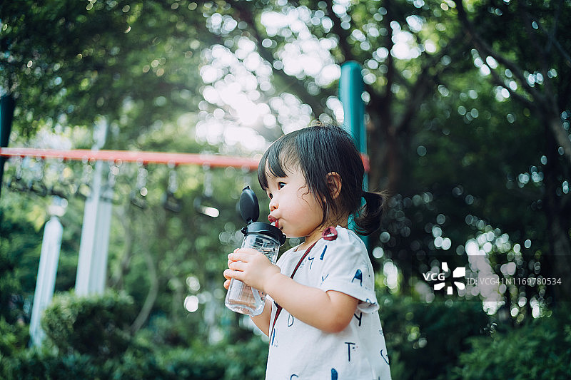 汗流浃背的蹒跚学步的小女孩在户外操场上玩耍后喝着水壶里的水图片素材