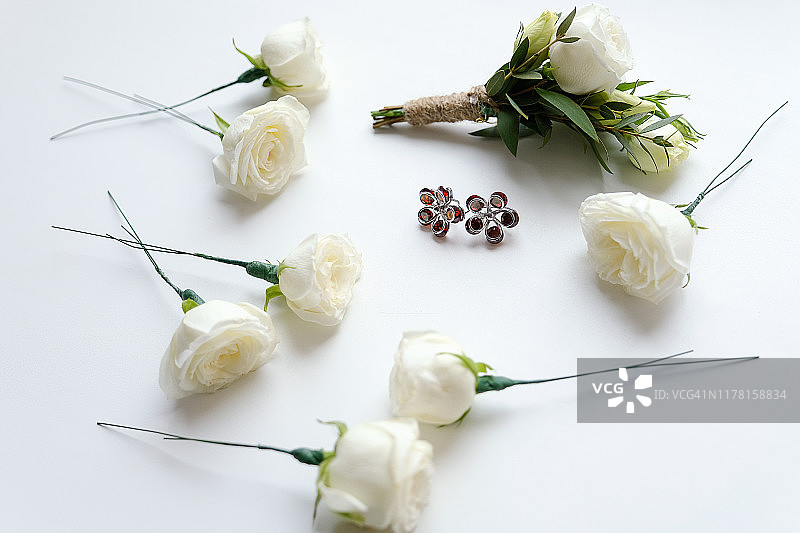 新郎的白玫瑰胸花和绿叶在白色的背景。靠近鲜花和耳环的新娘。婚礼用品和配件。图片素材