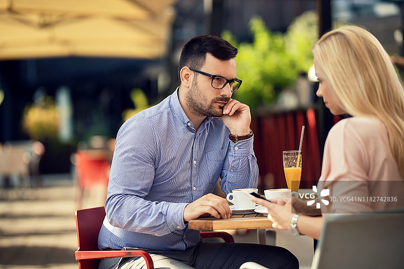 一个不高兴的男人看着他的女朋友，她正在一个咖啡馆用手机发短信。图片素材