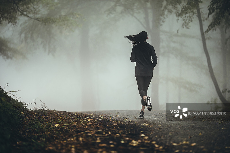 田径妇女在秋天的一天在雾蒙蒙的森林里跑步的背影。图片素材