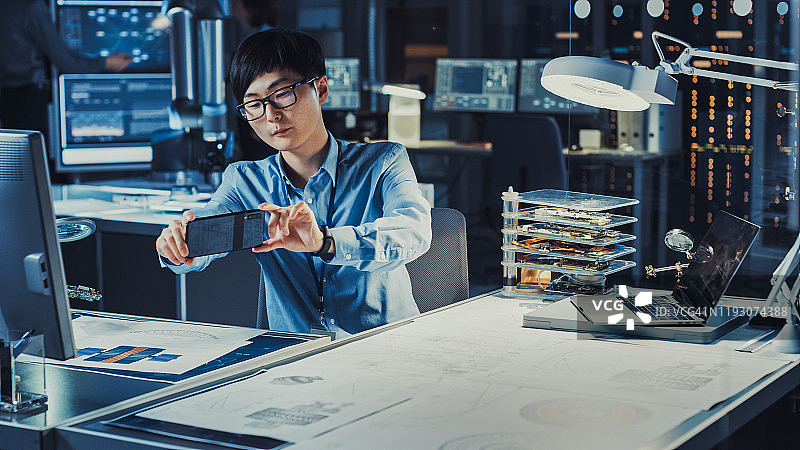 英俊的日本开发工程师穿着蓝色衬衫正在看增强现实技术图纸上的Smartpgone在高科技研究实验室与现代计算机设备。图片素材