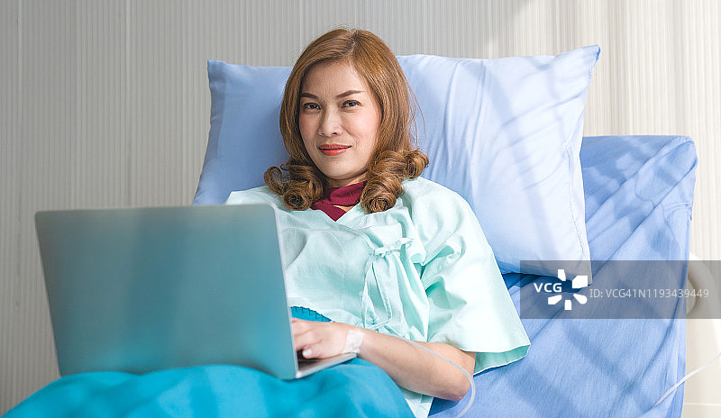 亚洲妇女生病的病人躺在医院的床上使用笔记本电脑和智能手机放松时，她的病在医院恢复。图片素材