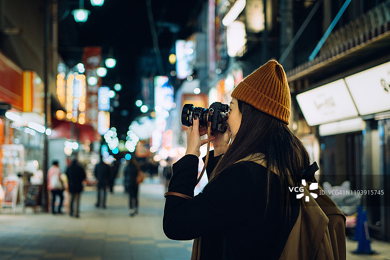 在日本大阪，年轻的亚洲女游客微笑着用相机拍照，一边探索一边漫步在繁忙的城市街道上图片素材