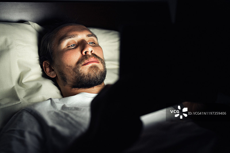 年轻英俊，蓄着胡子的疲惫男子在晚上看平板电脑图片素材