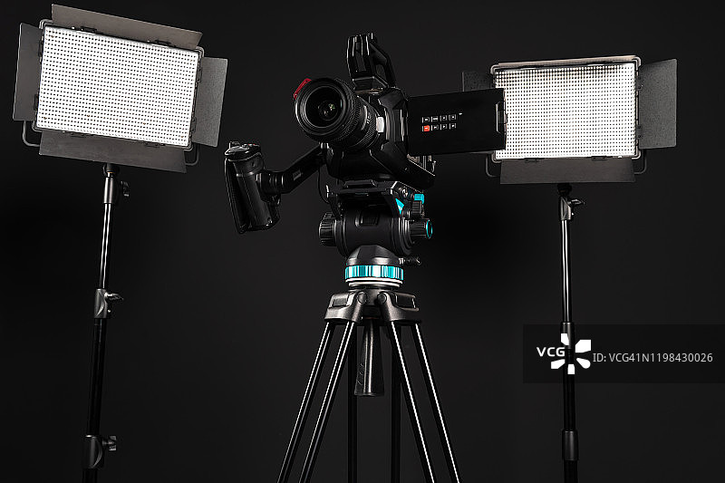 专业影院摄像机在一个摄像机三脚架和两个照明LED面板在黑色工作室背景图片素材