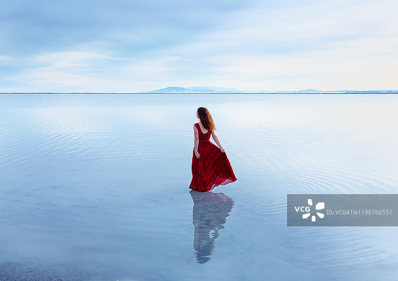 后视图的年轻女子与长波浪红头发在无袖红色连衣裙走在浅静止的湖图片素材