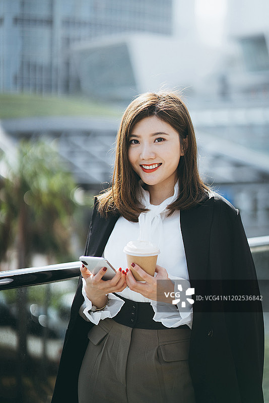 自信的年轻亚洲女商人在现代写字楼前用智能手机和咖啡对抗城市场景图片素材