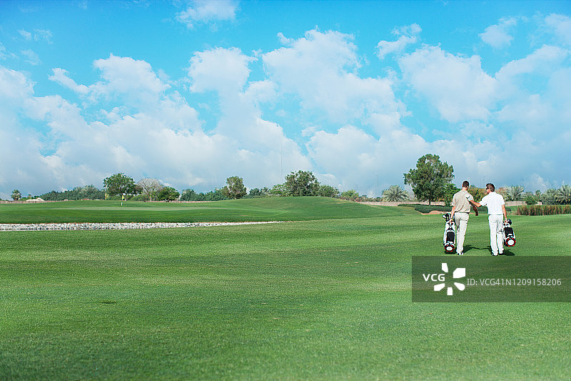 两名男子扛着高尔夫球袋，在高尔夫球场上行走。图片素材