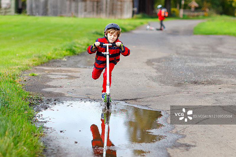 可爱的小男孩骑着滑板车在往返学校的路上。7岁的小男生开车穿过雨坑。有趣的快乐的孩子在五颜六色的时装和头盔。图片素材