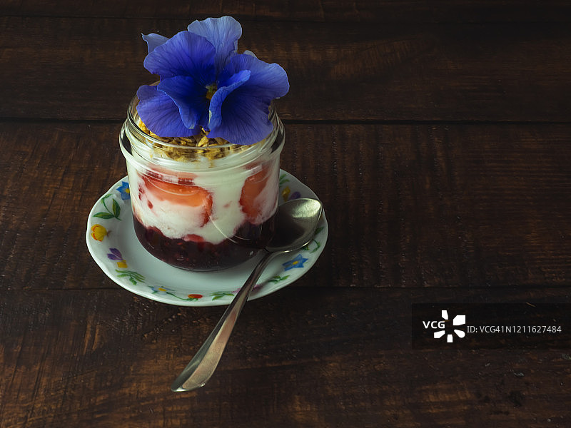 一杯加草莓、红莓果酱的原味酸奶和早餐格兰诺拉麦片，一朵可食用的花，放在木桌上的一把旧银勺或羊驼勺图片素材