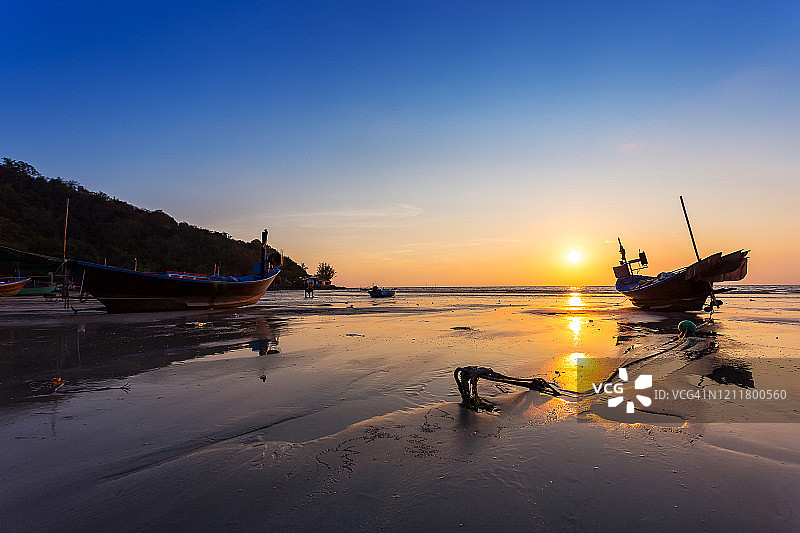 在泰国的日落时分，小渔船的锚躺在海滩上，水面的倒影和海滩上黄、橙的夕阳色图片素材