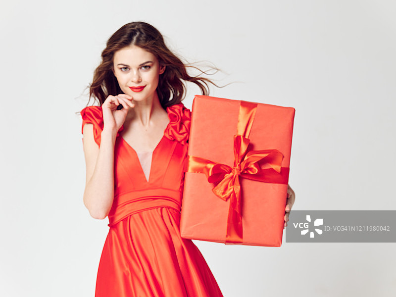 满意的年轻女孩穿着红色晚礼服正拿着她的大红色盒子和礼物。图片素材