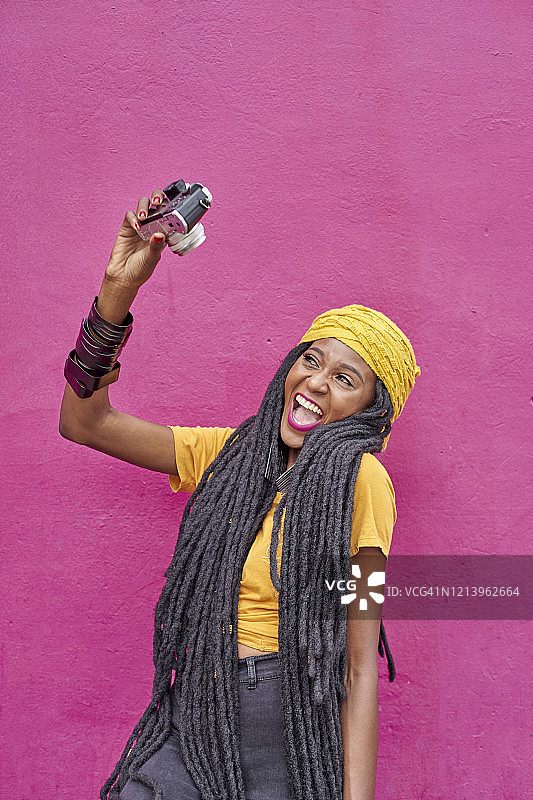 一个留着长辫的女人用她的相机在一堵粉红色的墙前自拍图片素材