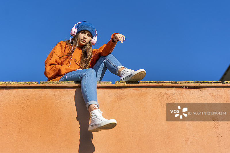 一个十几岁的女孩坐在墙上，背靠天空，戴着耳机听音乐图片素材