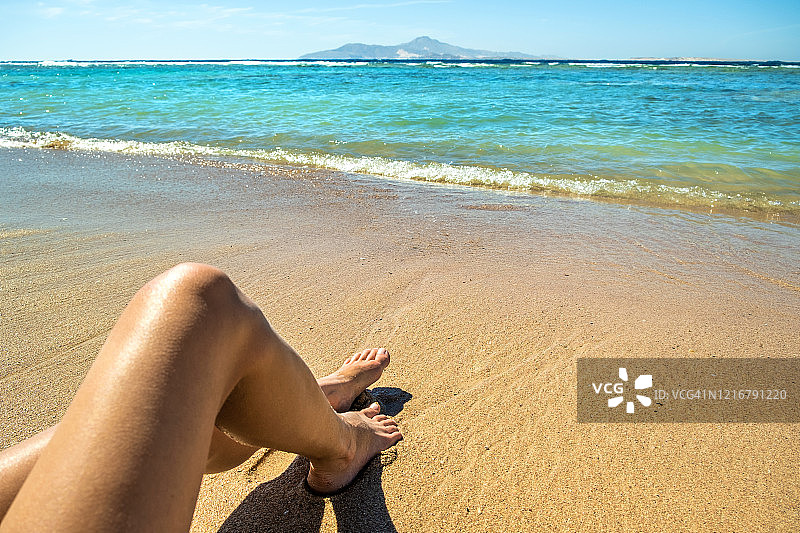 长而苗条的年轻女人的腿放松躺下和日光浴在沙滩上热带海滩在炎热的太阳在夏天。护肤、防晒、防老化和海上旅游理念。图片素材