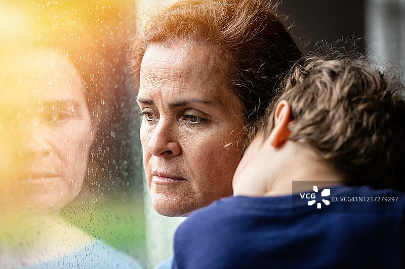 一名成熟女子和儿子合影，非常悲伤地透过窗户向外看，担心2019冠状病毒病被封锁图片素材
