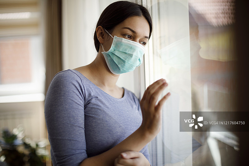 年轻悲伤的妇女站在窗前戴着医用防护口罩的肖像，以防止病毒和细菌在家里流行的疾病图片素材