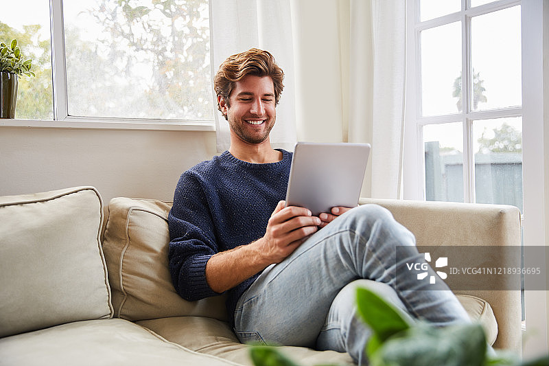微笑的年轻人在他的客厅沙发上使用数字平板电脑图片素材