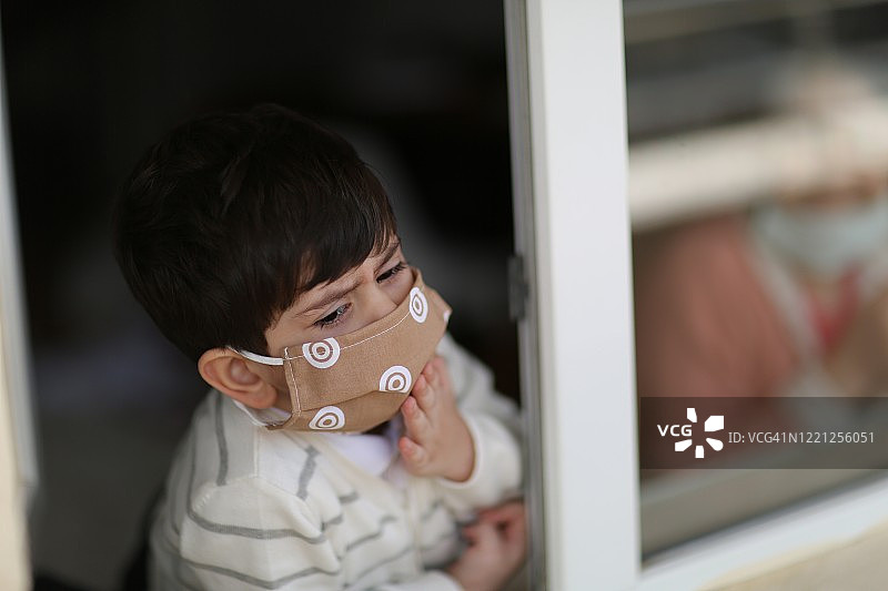 一个戴着医用口罩从窗户往外看的小男孩图片素材